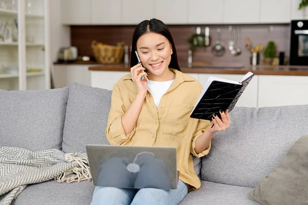 Multitask asian freelance woman talks on the smartphone and takes notes with pen, using laptop for remote work from home, write down some information sitting on the sofa. Girl works remotely