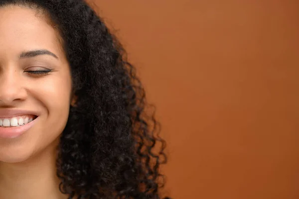 Half face portrait of charming multiracial lady laughing against brown wall. Young beautiful african-american woman isolated on brown background posing with closed eyes and smiles toothy
