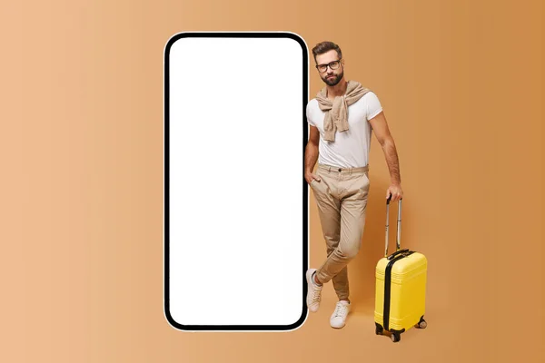 Travel app. Handsome charismatic bearded man stands with luggage near huge mobile phone with empty screen, modern hipster guy in glasses with yellow suitcase advertising app for booking and traveling