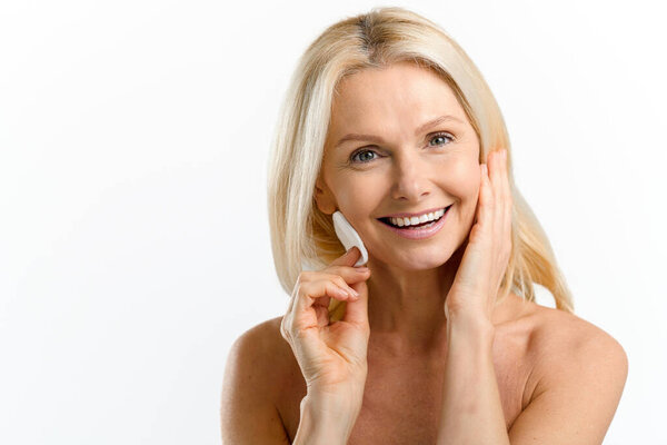Delighting mid age woman with smooth face skin applying lotion with cotton pad isolated on white. Charming 40s lady with naked shoulders removing make-up. Skincare routine
