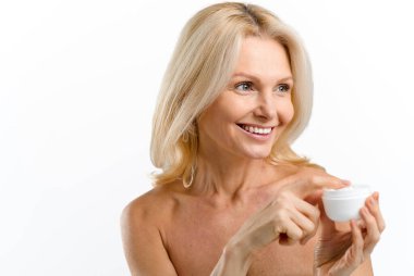 Beyaz arka planda izole edilmiş krema kavanozuyla poz verirken, yaşlı, çıplak, beyaz bir kadın başka tarafa bakıyor. Deri bakımı konsepti. Yeni kozmetoloji ürünü