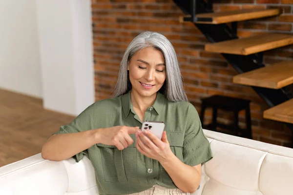 Calm senior mature lady sitting in a relaxed pose on the comfortable couch with a smartphone, woman spends leisure time scrolling news feed, web surfing