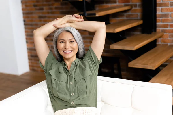 Positive middle-aged woman with long hair at the sofa in modern apartment. 50s asian female resting at home, looks at the camera. Wellbeing concept