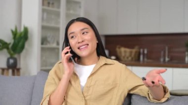 Gülümseyen genç bir kadın cep telefonuyla arkadaşlarıyla sohbet ediyor, keyifli telefon konuşmaları yapıyor. Çekici Asyalı kadın akıllı telefonda konuşuyor evdeki rahat koltukta oturuyor.