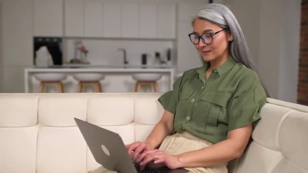 ソファに座って プロジェクトに取り組んで快適なカジュアル服でシニアアジアの女性ビジネス女性やフリーランス 喜びの笑顔で画面を見て — ストック動画