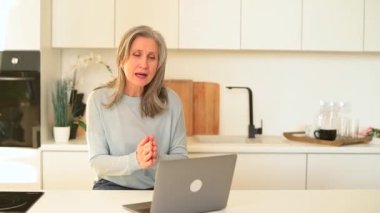 Yakışıklı, olgun bir kadın video bağlantısı için dizüstü bilgisayar kullanıyor, arama yapıyor, bilgisayar ekranına bakıyor ve el sallıyor. Gri saçlı büyükanne ailesiyle irtibat halinde.
