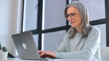 Gözlük takan şık, olgun bir iş kadını modern ofis alanındaki masada dizüstü bilgisayarını kullanıyor. Kendinden emin, gri saçlı, e-postaları cevaplayan, rapor hazırlayan, araştırma yapan yaşlı bir bayan.
