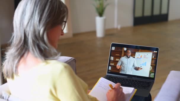 オンライン語学コースを受講する上級女子学生 アフリカ系アメリカ人の教師がオンラインのウェビナーを行っている間 ノートを取りながら ビデオクラスを見るためにラップトップコンピュータを使用して成熟した女性 バックビュー — ストック動画
