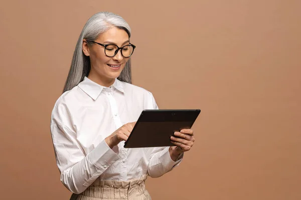 Modern elderly senior woman in formal wear using digital tablet isolated on beige. Portrait of mature female business person using online technology, computer app