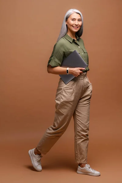 Positive smiling woman with grey hair standing with closed laptop in hands, while having break. Indoor studio shot isolated on beige background