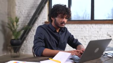 Front view of positive Hindi man in smart casual shirt using laptop while sitting at the desk in his flat. Young Indian male student watching webinars, educational courses, learning on the distance