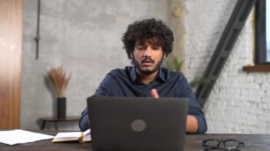 Video iletişimi için dizüstü bilgisayar kullanan neşeli, çekici Hintli adam. Modern ofisteki masada oturan gözlüklü adam webcam 'e el sallıyor ve internette konuşuyor.