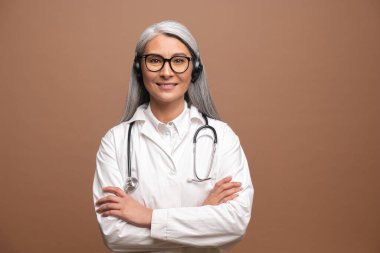 Steteskoplu beyaz elbiseli, orta yaşlı Asyalı kadın doktor hastayı aramak için kablosuz kulaklık kullanıyor, kahverengi üzerine izole edilmiş kameraya bakıyor. İnternette danışmanlık ve terapi