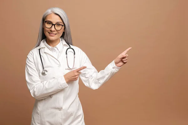 Wow look here. Portrait of happy middle-aged 50s Asian woman doctor pointing aside at empty place, standing isolated on brown background, copy space for promotional ad