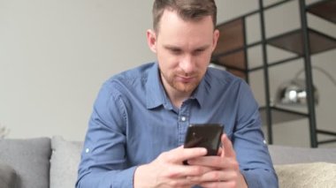 Rahat bir oturma odasındaki kanepede oturan ve elinde akıllı telefon tutan sakin genç adam, erkek arkadaşlarıyla mesajlaşıyor, sosyal ağlarda haber yayını yapıyor.