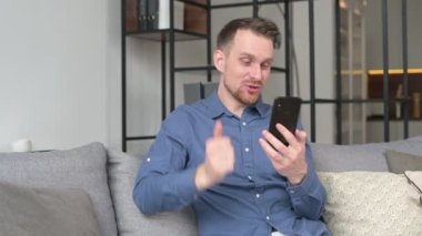 Genç adam video görüşmesi için akıllı telefonu kullanıyor evdeki kanepede oturuyor, elinde telefonu tutuyor, el sallıyor merhaba, video görüşmesi, sanal görüşme.