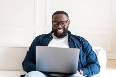 Tişört ve kot pantolon üzerinde gülümseyen Afro-Amerikan adam modern dairede koltukta oturuyor, siyahi adam serbest çalışıyor, girişimci, erkek öğrenci dizüstü bilgisayar kullanıyor, internette sohbet etmekten hoşlanıyor.