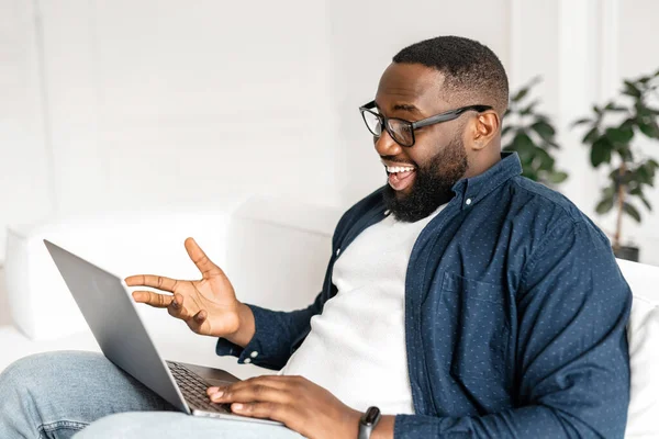 Excited happy 30s African guy enjoying virtual meeting, cheerful glad black man making video call on the laptop, sitting on the sofa at home, stay in touch with friends or family on the distance