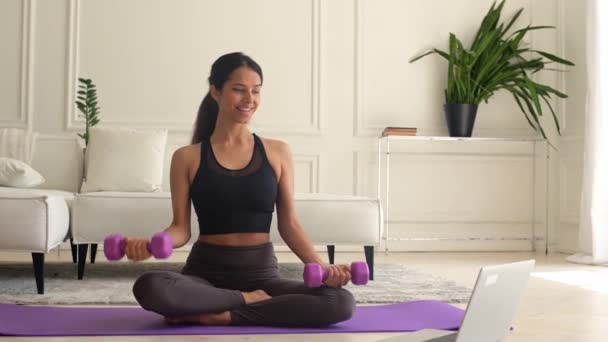 Healthy lifestyle concept. Cheerful young fit indian woman is doing exercises on the mat at home and watching video tutorial on laptop, exercising at home, using dumbbells to swing arm muscles