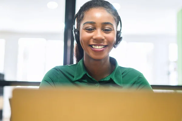 Smiling african-american female employee colleague using headset and laptop sitting on the workplace, talking on the distance, work in call center, multiracial woman is customer support operator