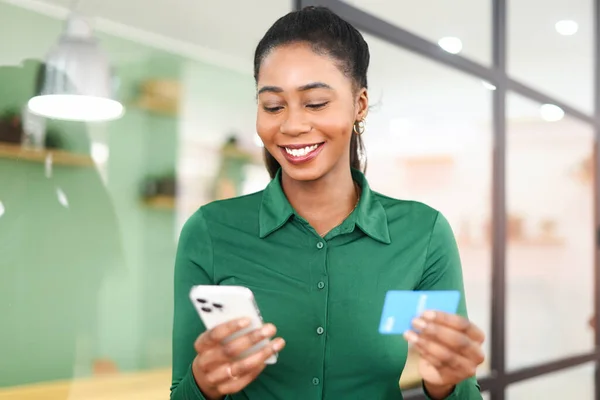 Cheerful african-american young woman holding smartphone and credit card, making online money transaction standing with the office background, online shopping, e-banking