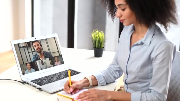 聪明的混血女商人坐在办公桌前 通过笔记本电脑与一群不同的同事通过视频通话 进行虚拟在线会议 做笔记 用记事本书写 — 图库视频影像