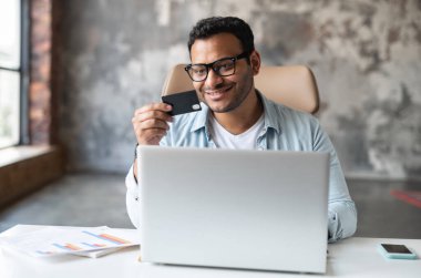 Gözlüklü başarılı Hintli işadamı, gülümsüyor, dizüstü bilgisayarın başında oturuyor, internetten ödeme yapıyor, internetten satın alıyor, ofisten çalışıyor, kredi kartı tutuyor, internetten alışveriş yapıyor,