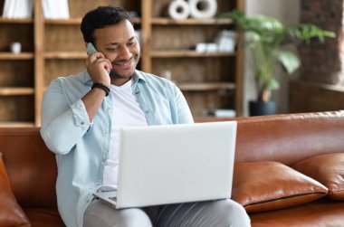 Serbest giyinen Hintli adam bilgisayarla uzaktan kumandalı iş yapıyor ve akıllı telefonla konuşuyor. Melez adam telefonla konuşuyor ve evdeki kanepede oturuyor.