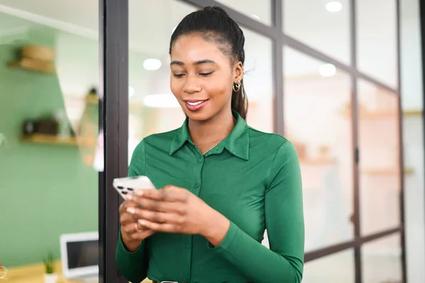 Smiling African-American female employee using modern smartphone, texting, messaging via wireless internet connection, smiling african multiracial businesswoman browsing smartphone in modern office