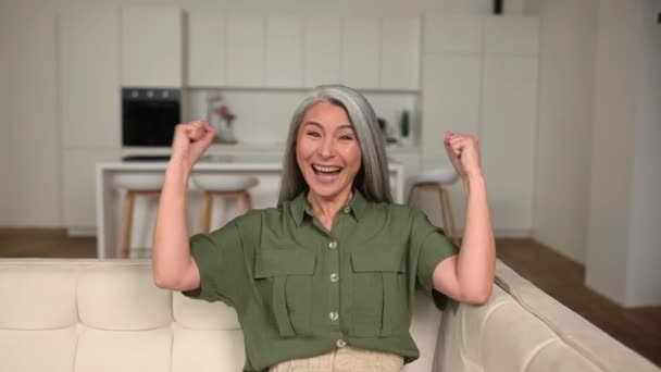 幸运的是 资深亚洲女人庆祝胜利 兴奋的 成熟的 灰白头发的女士坐在家里的沙发上 举起拳头表示胜利 她得到了好消息 — 图库视频影像
