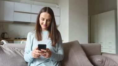 Serene genç kadın akıllı telefonu evdeki kanepede oturmuş arkadaşlarıyla sohbet ediyor, boş zamanlarını sosyal medyada geçiriyor, mesajlaşıyor, haber yayını yapıyor, internetten alışveriş yapıyor.