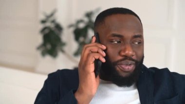 Neşeli ve gülümseyen Afro-Amerikan bir adamın yakın plan portresi. Evde akıllı telefonda sohbet ediyor. Melez sakallı adam, telefon konuşmasından memnun.