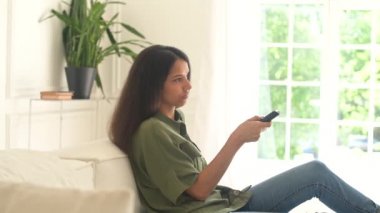 Neşeli genç Hintli kadın evdeki rahat kanepeye oturur ve uzaktan kumandayı elinde tutar, televizyon izler ve tek başına dinlenir, boş zamanlarını dizileri izleyerek geçirir, televizyon programlarından zevk alır.