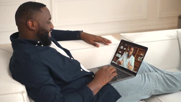 非裔美国人可以与朋友 男同事或老师进行虚拟会面 多种族人士正在使用笔记本电脑进行视频通话 躺在沙发上轻松交谈 与非洲男人交谈 — 图库视频影像