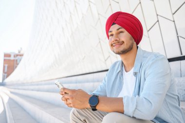 Ulusal başörtülü Hintli adam merdivenlerde oturuyor, akıllı telefon tutuyor, sosyal ağlarda vakit geçiriyor, mesajlaşıyor ve sohbet ediyor, rüya gibi bir şekilde yüzünü çeviriyor.