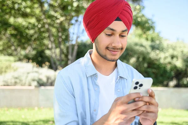 Close-up portrait of smiling young Indian man wearing red turban pagg texting on the smartphone, using mobile phone for communication online, hindu male freelancer developer testing app on the phone