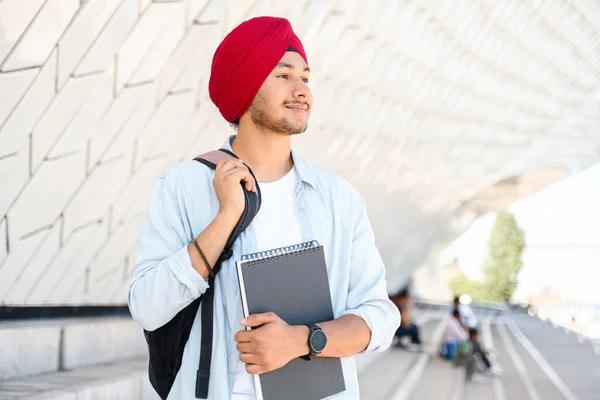 Inspired Indian male student with backpack holding laptop outdoors, foreigner hindu guy proud to be university member, hindu guy in traditional turban looking aside standing outdoors happily