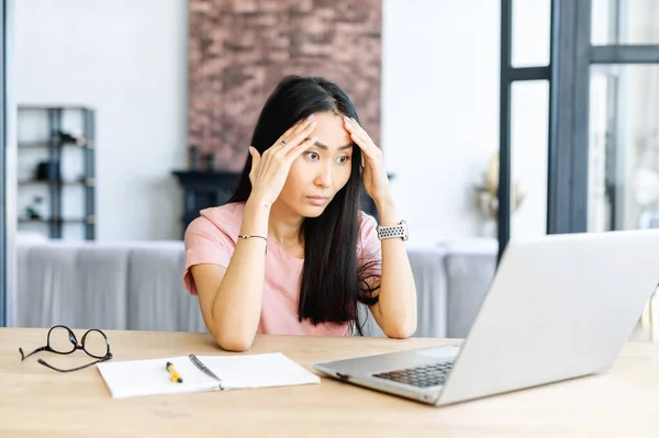 Troubled and frustrated young Asian female office worker holding head in hands, sitting alone at the modern office desk with an open laptop, stressing out, cannot fix a problem, staring at the screen