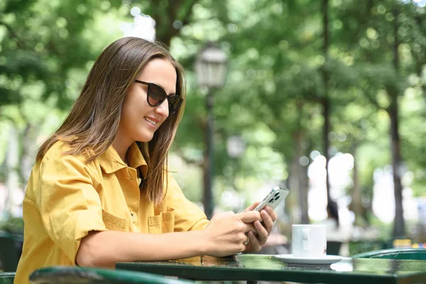 在户外用智能手机观看微笑的年轻女性 在暑期咖啡店的咖啡休息时间上网发短信 戴着太阳镜和黄色休闲衫的积极微笑的女性在社交网络中度过时光 — 图库照片