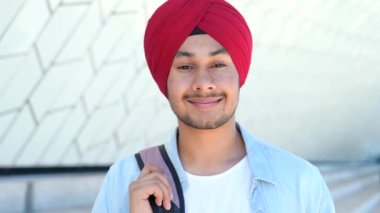 Sırt çantalı, kırmızı milli başlıklı modern Hintli öğrenci dışarıda dikiliyor, Hintli adam hoş bir gülümsemeyle kameraya bakıyor.