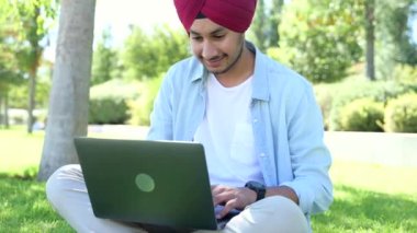 Erkek Hintli öğrenci geleneksel kırmızı başlıklı türban giyiyor çimenlerde dizüstü bilgisayar kullanıyor, kampüsün yakınlarında ödev hazırlıyor, Hintli adam daktilo ediyor, e-postalarına cevap veriyor.