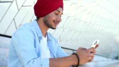 Portrait of cheerful young Indian guy in turban sending message on mobile phone sitting on the steps outdoors. Happy ethnic man using smartphone, enjoying chatting online, messaging in social networks