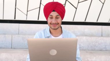 Cheerful Indian man wearing headwrap sitting on the steps outdoors and using laptop. Inspired glad male entrepreneur typing on keyboard and looks at the screen with pleasure smile