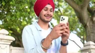 Carefree young Indian guy in casual shirt and traditional headdress using smartphone outdoors. Male student texting with classmates online, scrolling news feed in social networks, checking schedule