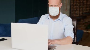Concentrated redhead bearded man wearing protective medical mask using laptop computer sitting in contemporary office space