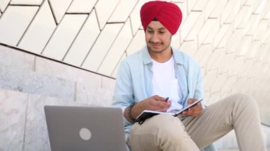Indian student man in traditional headwrap sitting outdoors and using laptop for learning on the distance, hindu guy watching webinar and taking notes, male student studying online, e-learning