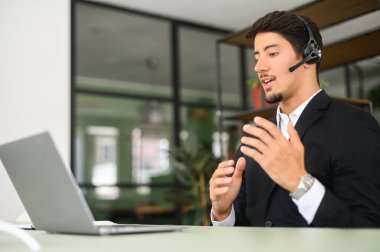 Kulaklık takan yardımsever ve dost canlısı İspanyol erkek çalışan ofiste dizüstü bilgisayarın önünde oturur, web kamerasına bakar ve uzaktan konuşur, iş adamı mikrofonlu kulaklıklar kullanır.