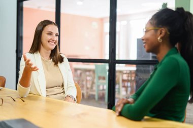 Two young happy multiracial female colleague sitting at desk and communicating while working together in coworking space, sharing new ideas, discussing project strategy