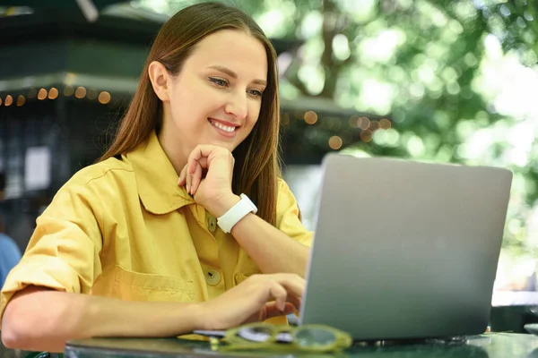 Cheerful female freelancer using laptop outdoors, enjoying remote work sitting at the table in summer cafe outdoors, smiling woman using laptop sitting in the park