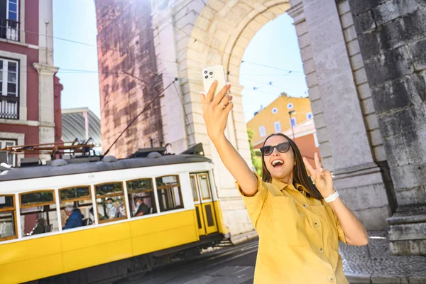 Overjoyed happy foreign woman tourist taking selfie with famous yellow tram, walking in old town of Lisbon, Portugal